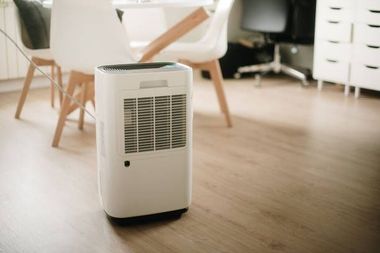 A white portable dehumidifier stands on a light wooden floor in a home office or dining room setting.