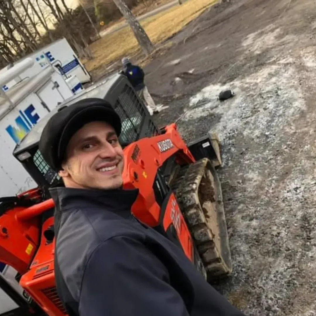A smiling person wearing a dark cap and jacket stands in front of an orange skid-steer loader at a construction site.
