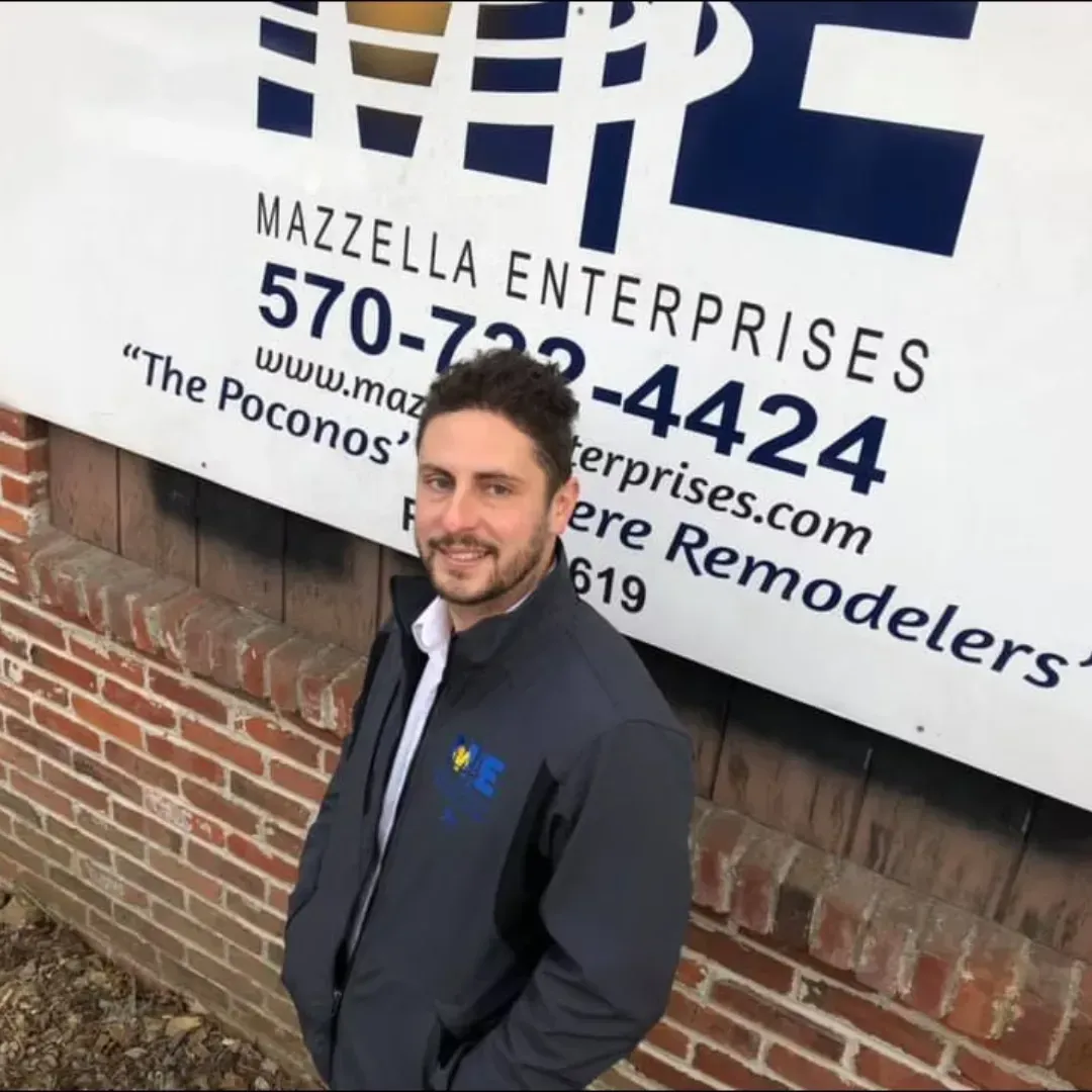 A person stands smiling in front of a sign for Mazzella Enterprises, featuring a phone number and company information.