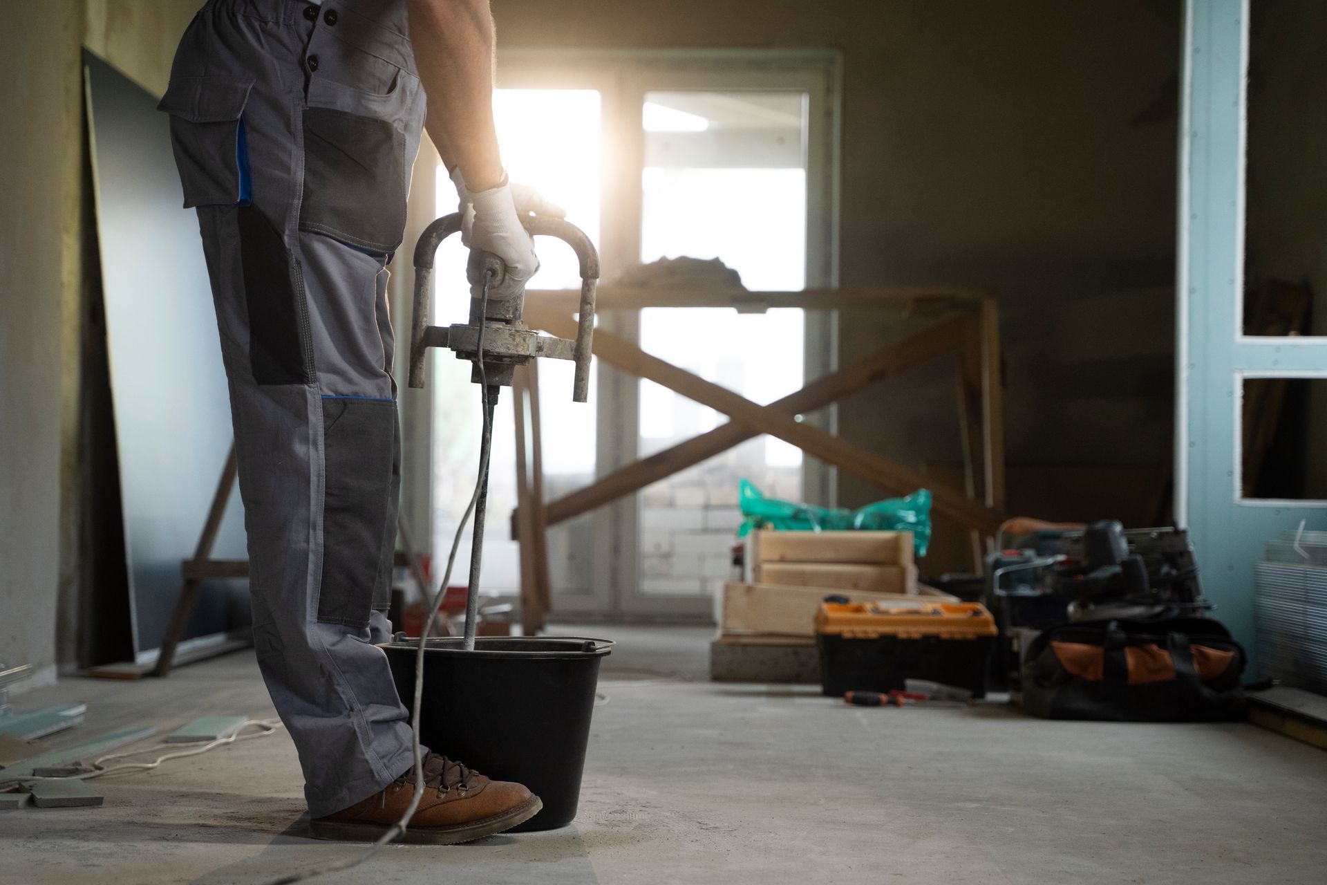 A worker in grey workwear mixes materials in a bucket with a handheld power drill in a dimly lit construction site.