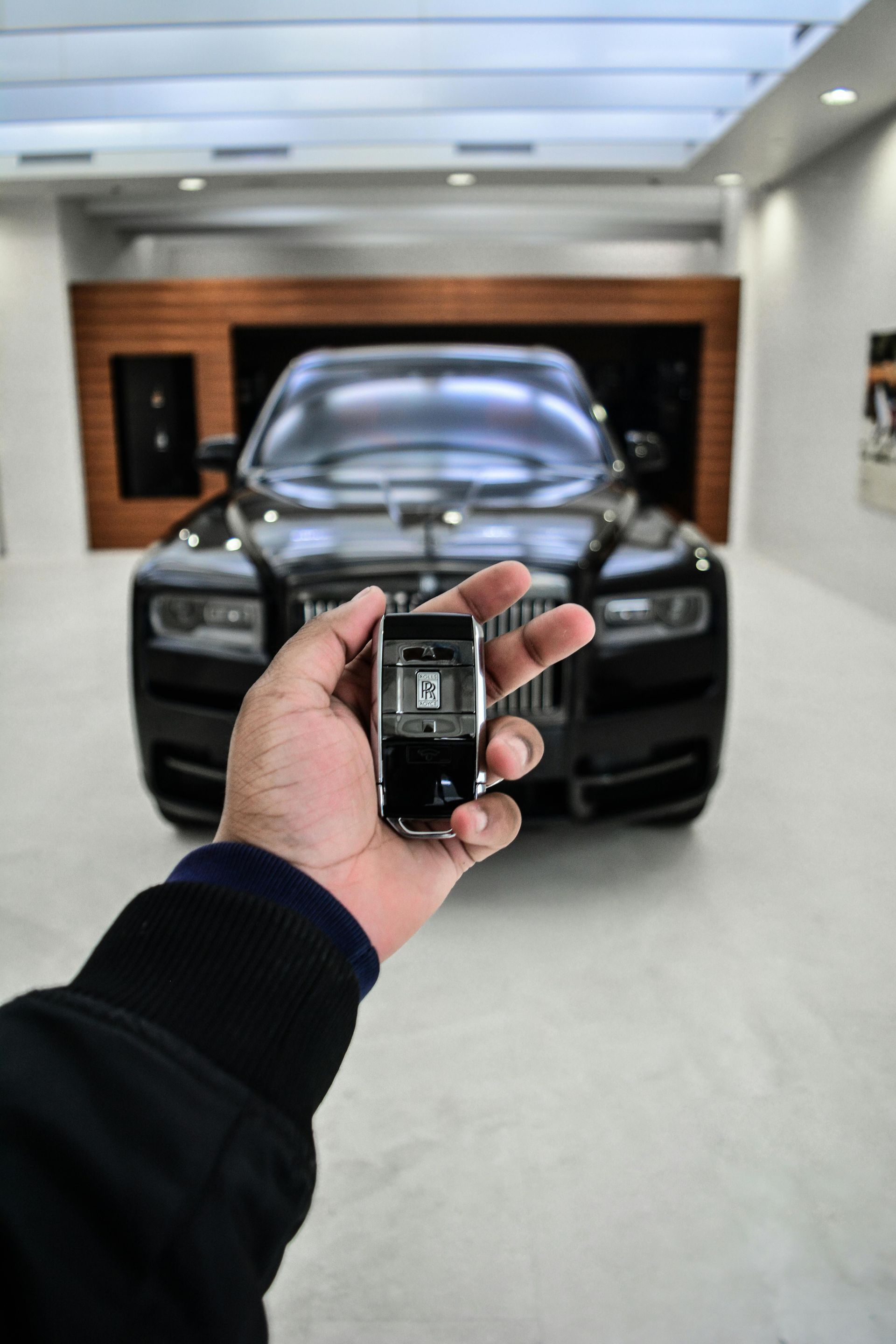 Person holding a Rolls-Royce key fob, a black Rolls-Royce car in the background, showroom setting.
