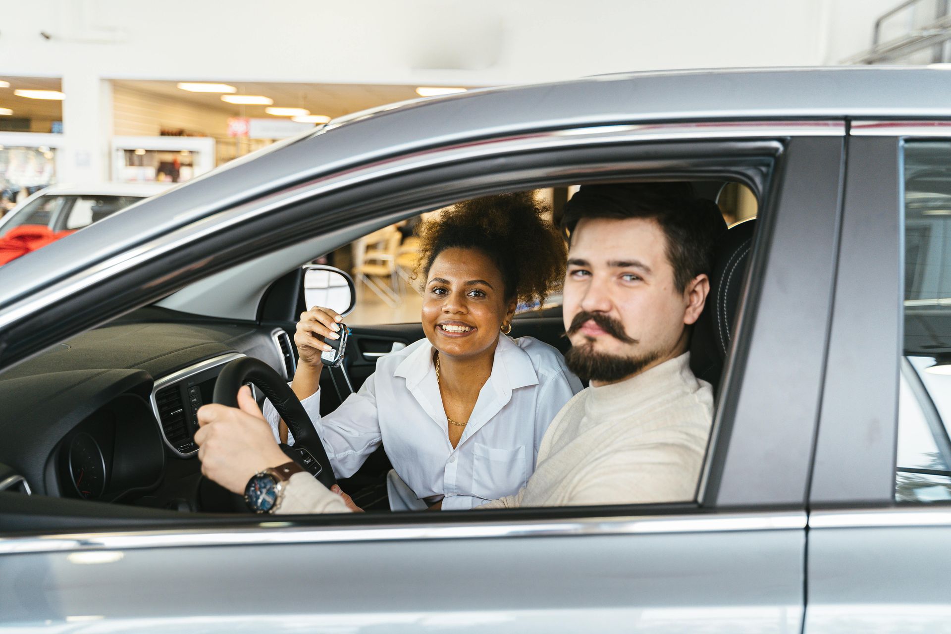 Woman holding glass, man driving a car, both smiling inside a car dealership.