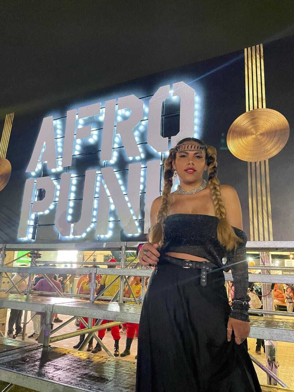 Mulher de vestido preto posando em frente a um letreiro iluminado com a inscrição “AFRO PUNK” à noite.