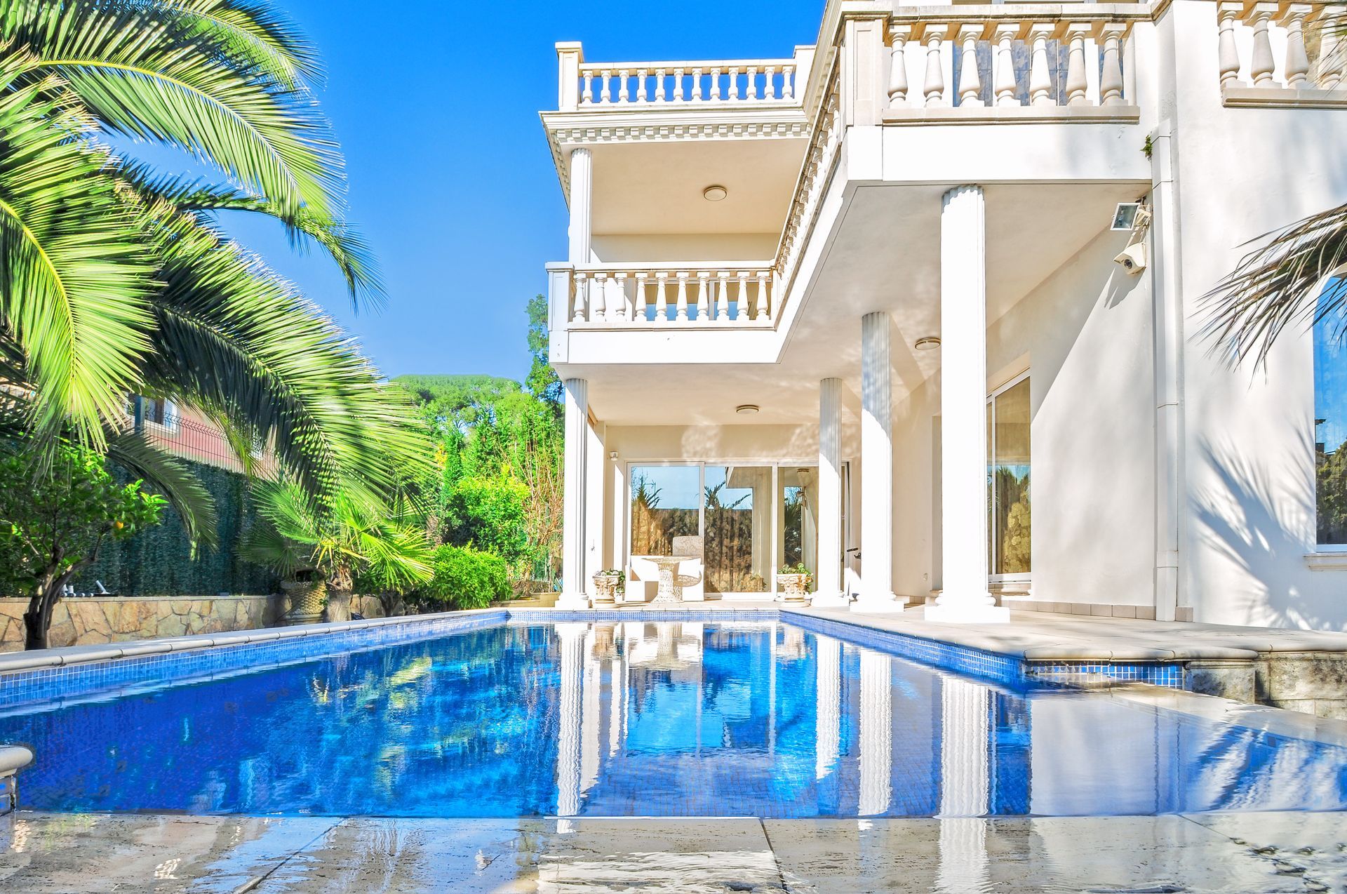 Luxury villa with a blue swimming pool, white columns, and palm trees under a bright sky