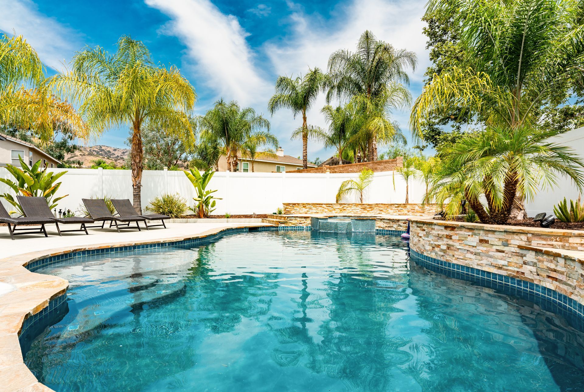 Backyard pool with palm trees, lounge chairs, and a stone waterfall under a blue sky