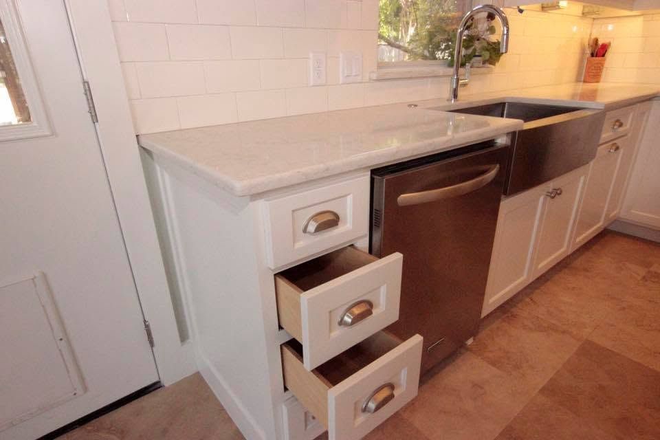 Kitchen cabinet with three open drawers beside a sink and dishwasher in a bright white kitchen
