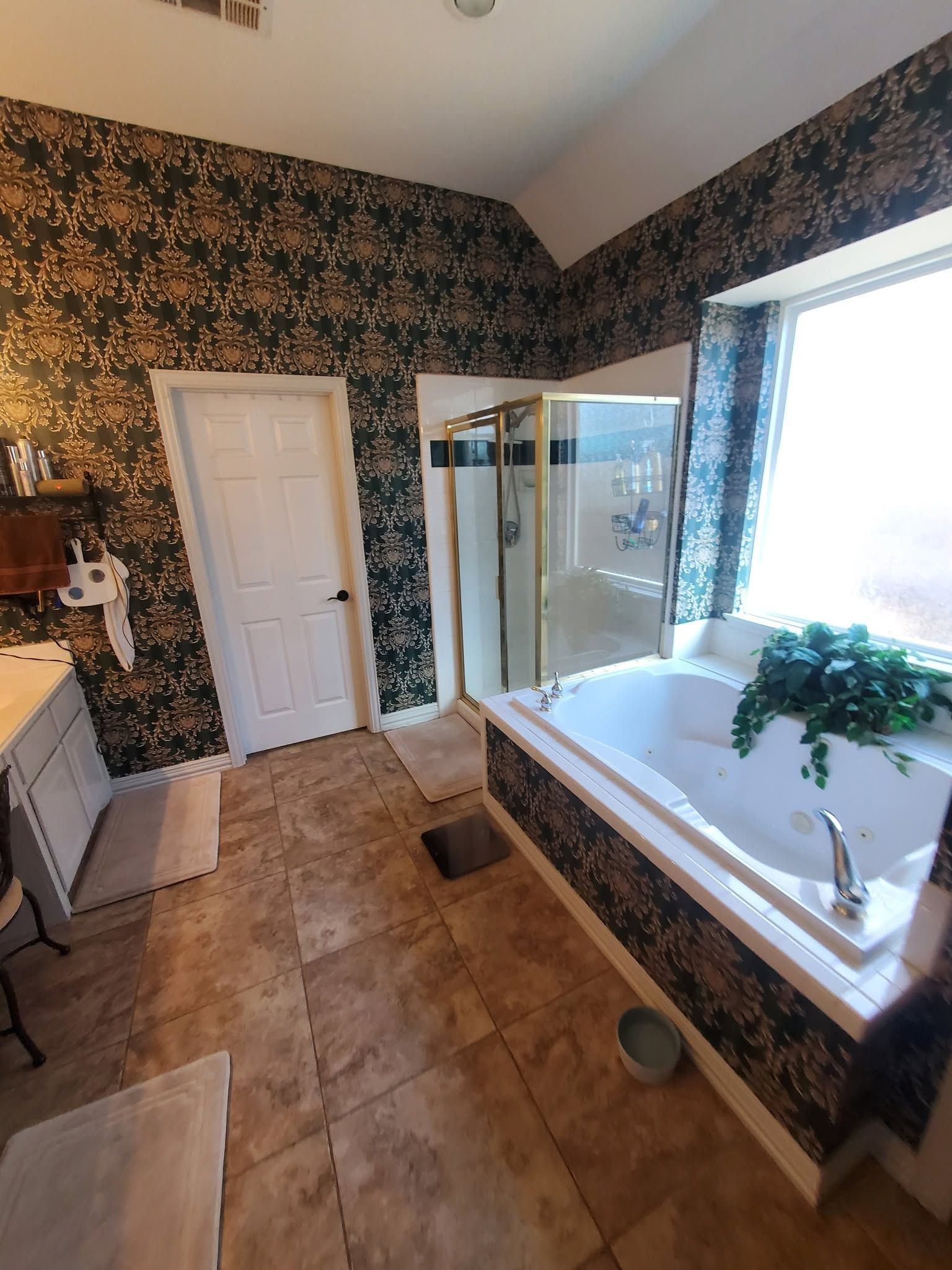 Bathroom with floral wallpaper, white tub, glass shower, and beige tile floor by a bright window