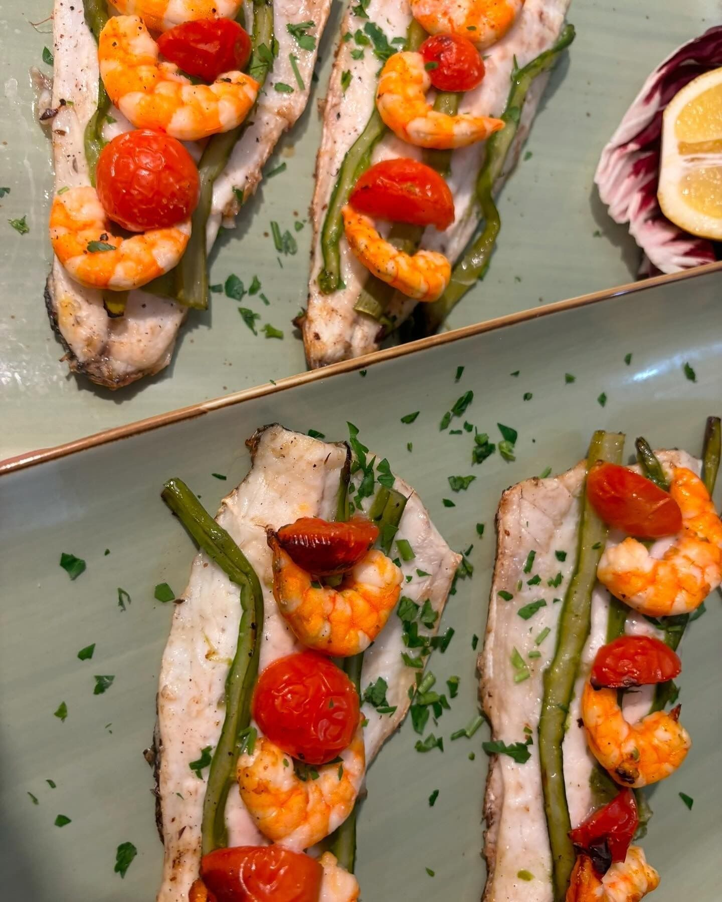 Filetti di pesce con gamberi, pomodori e asparagi su un piatto verde, guarnito con prezzemolo.