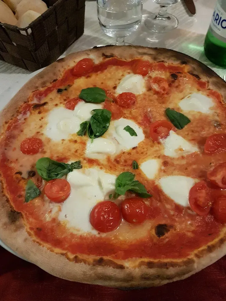Pizza con mozzarella, pomodorini e basilico su un tavolo apparecchiato con tovaglie rosse; sullo sfondo pane e acqua.