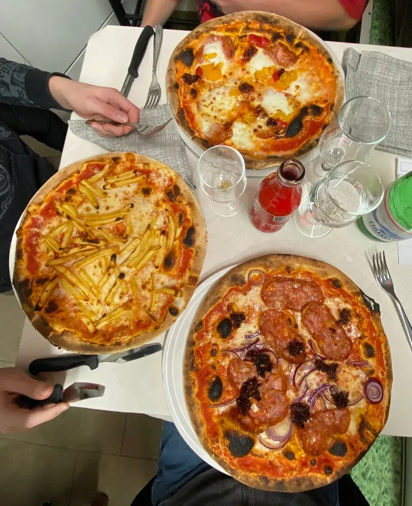 Tre pizze su un tavolo: una con le uova, una con le patatine fritte, una con il salame piccante. C'è gente presente.
