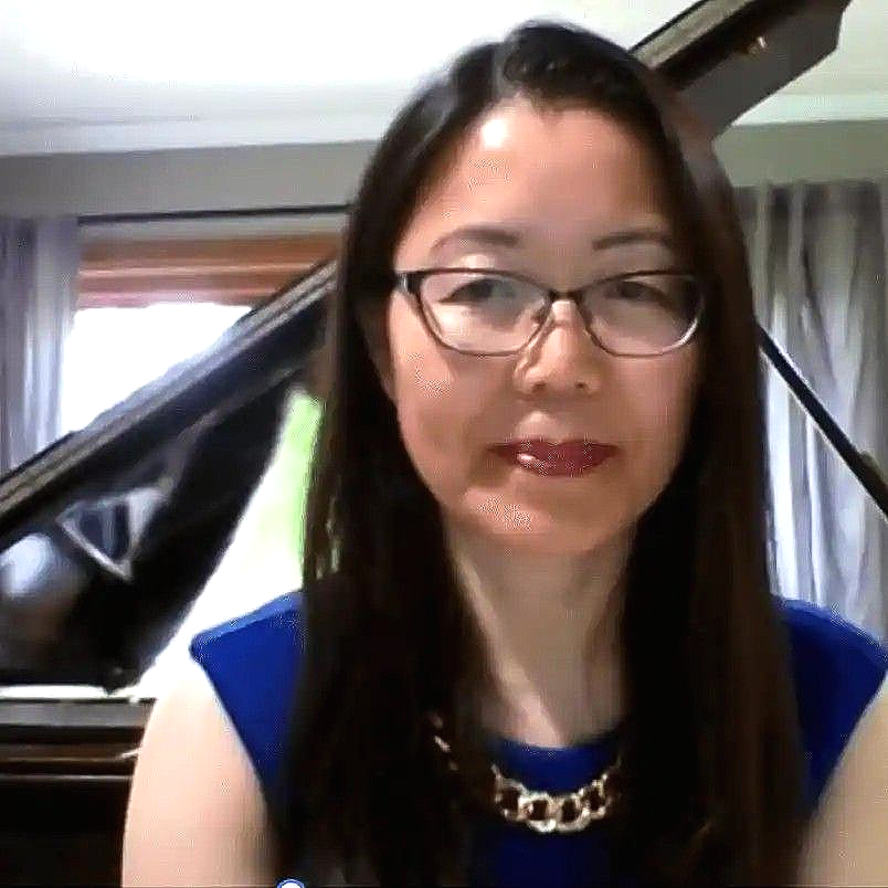 Woman in glasses and blue top seated indoors beside a grand piano