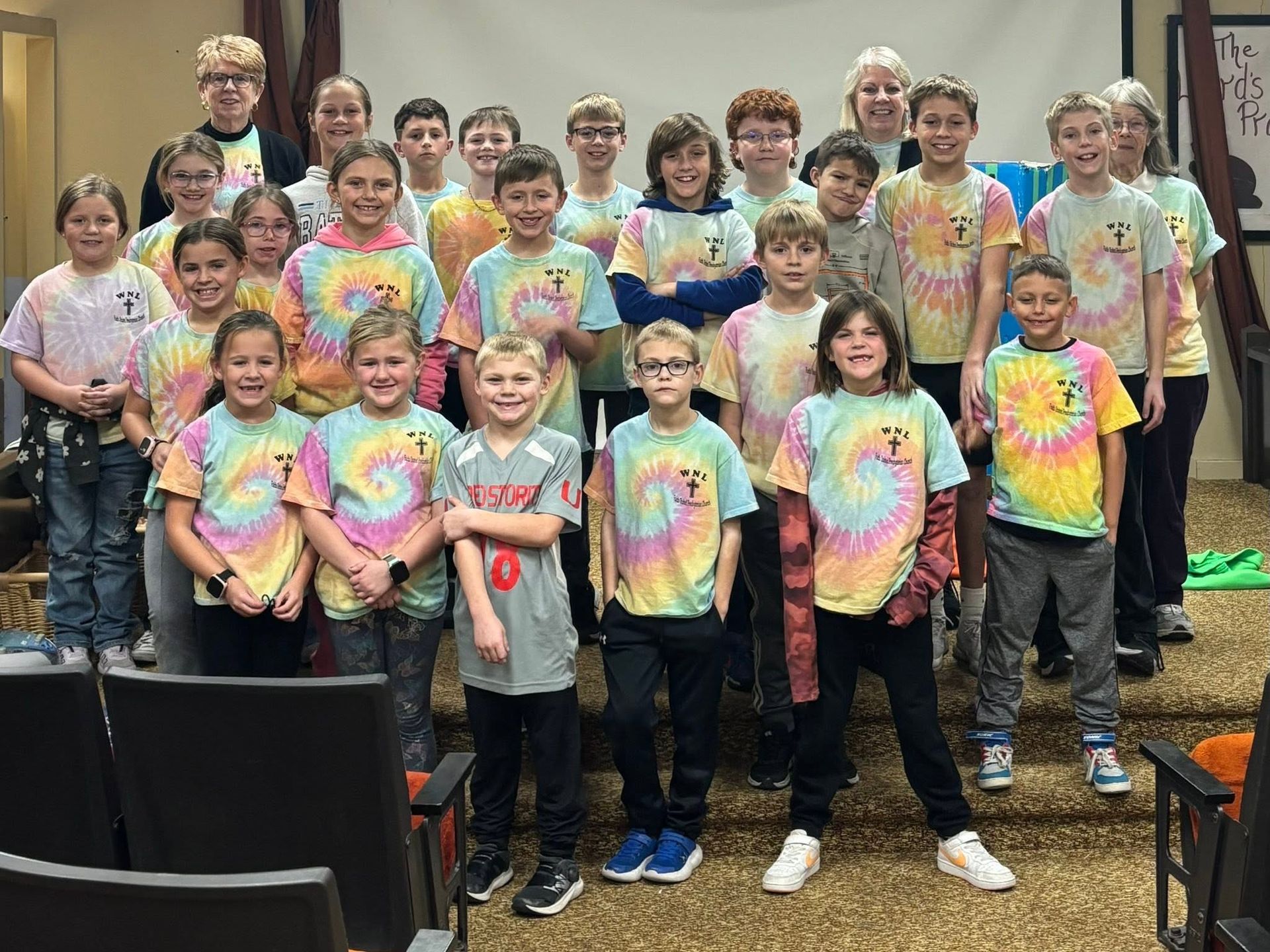 Group of children and adults in colorful tie-dye shirts posing together indoors on risers