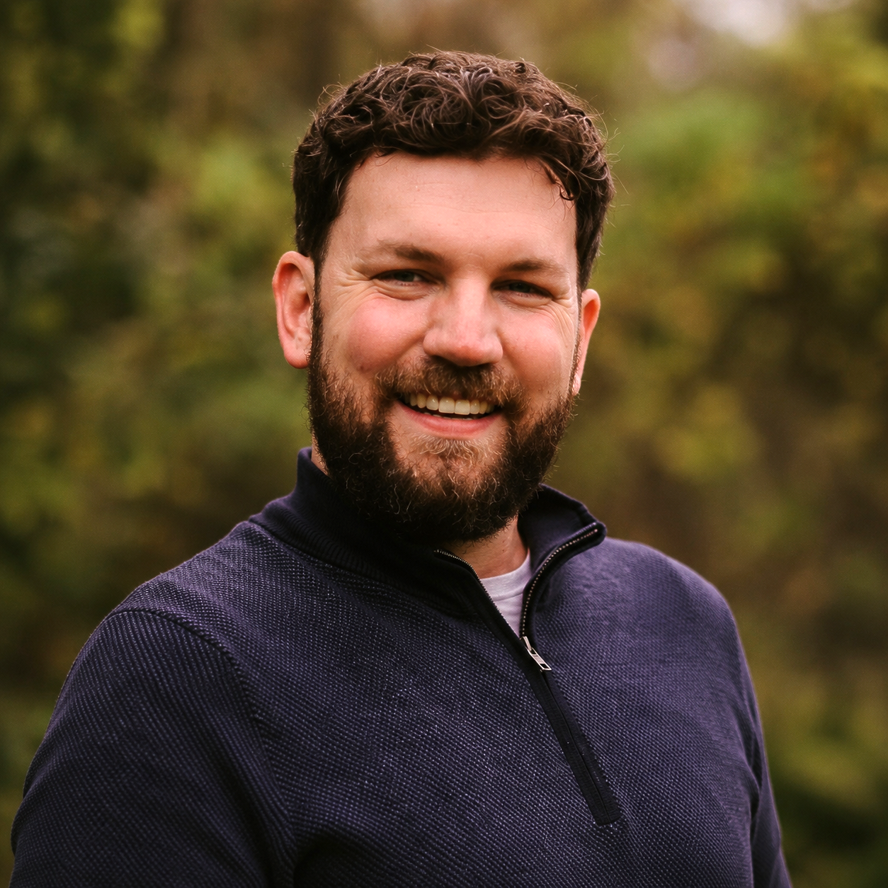 Smiling bearded man in a dark sweater outdoors against a blurred green background