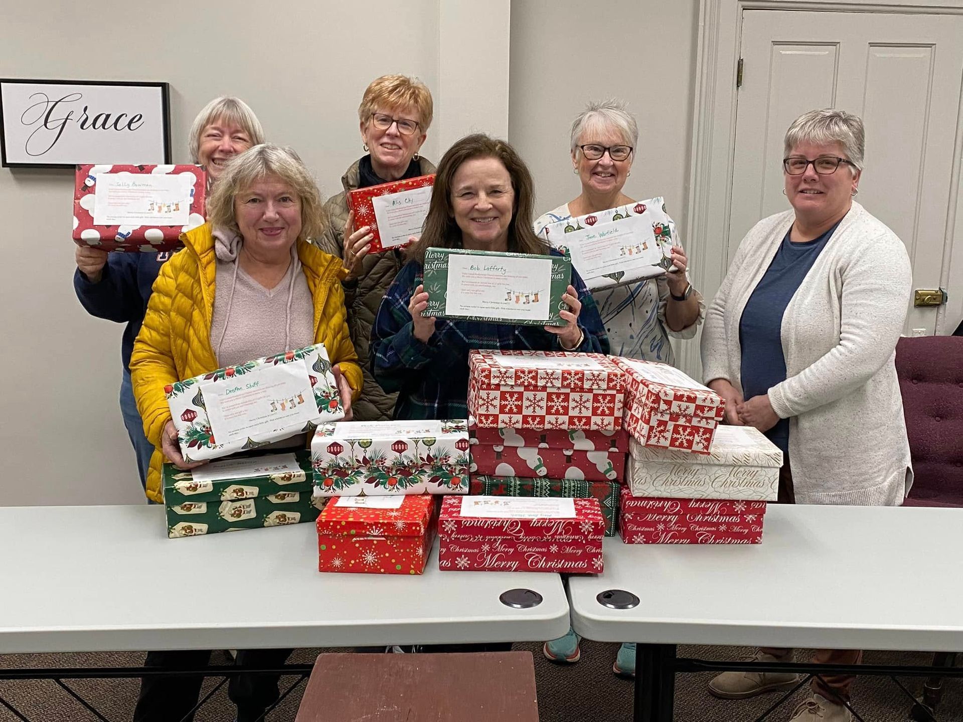 People posing with wrapped gift boxes and signs, smiling in a room at a table.