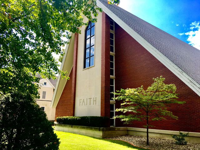 Modern church building with red brick, tall windows, and a steep roof beside trees and lawn