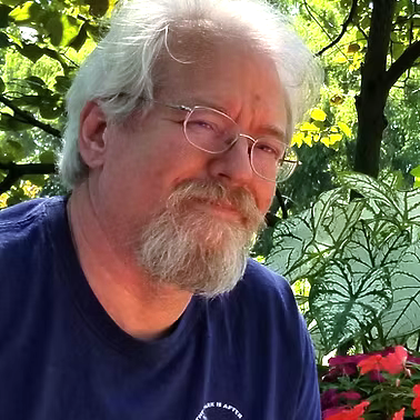 White-haired man with glasses and a beard smiling outdoors among trees and flowers