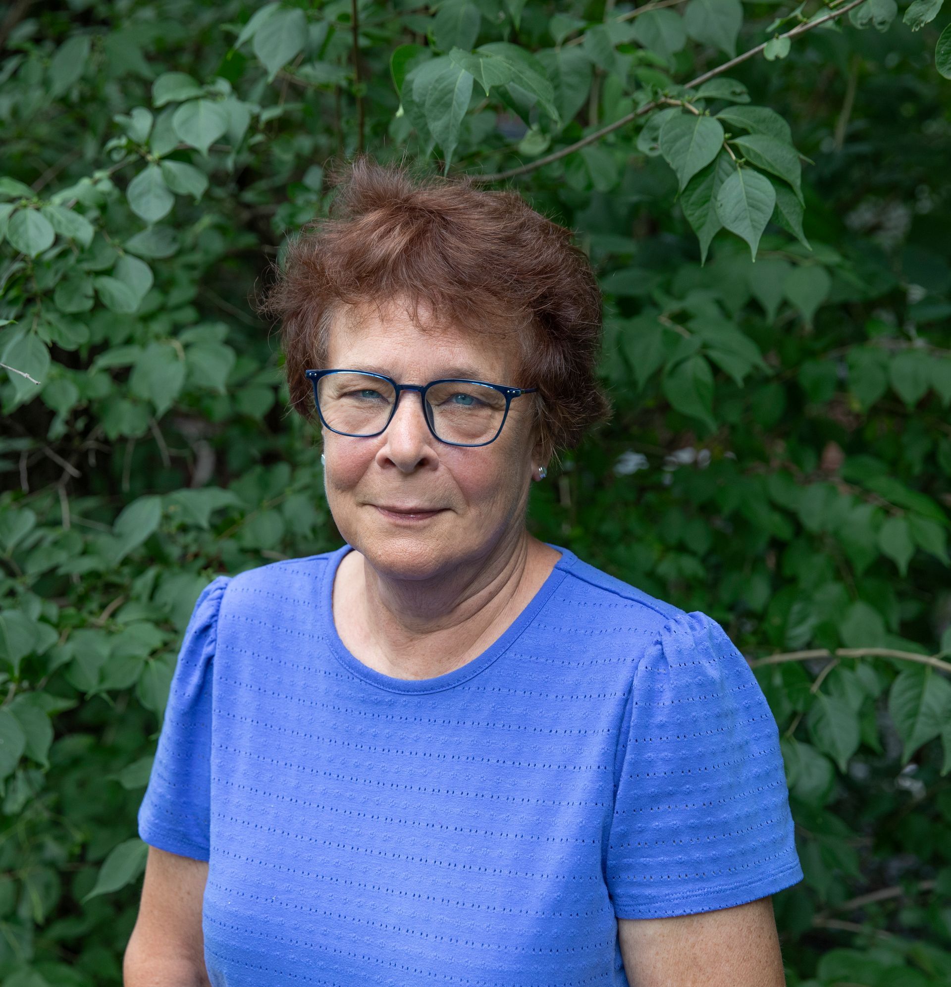 Woman in blue shirt and glasses standing before leafy green foliage