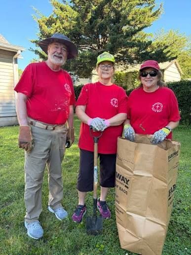 Three volunteers in red shirts stand outdoors holding gloves and a paper yard-waste bag.