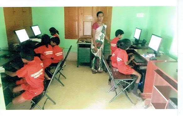 Computer lab with students in red shirts using desktop computers while an instructor stands nearby