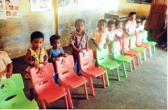 Children sitting on colorful plastic chairs in a classroom, facing forward in a row.