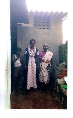 Three people standing outside a small house, wearing patterned clothes and smiling.