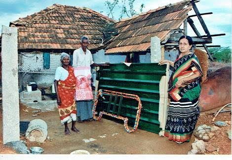 Three people stand beside a green cart outside a weathered house with a tiled roof.