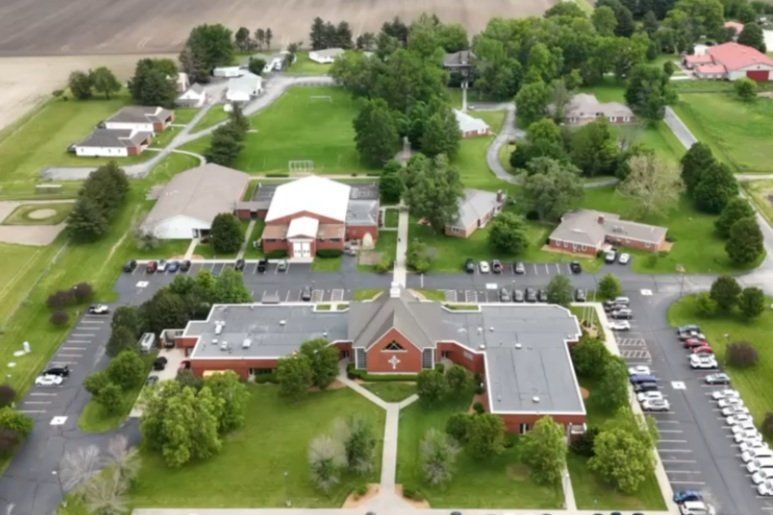 Aerial view of a large brick building with parking lots, lawns, and surrounding trees in a rural area