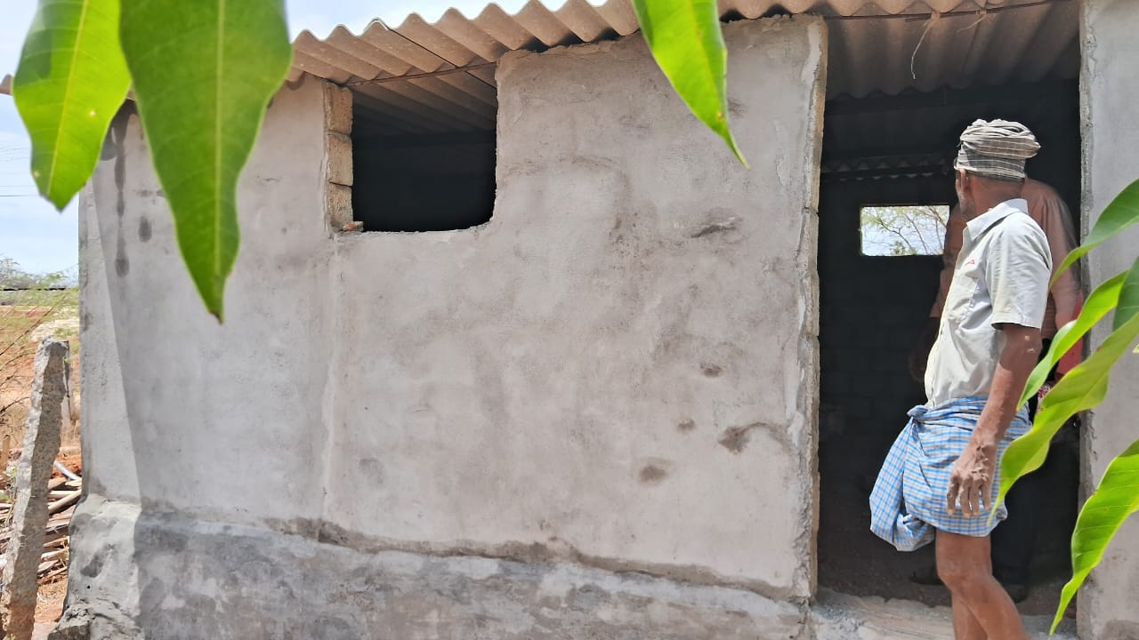 Small cement house with corrugated roof; person stands by the doorway holding a blue bundle.