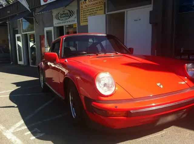 Red Porsche parked outside a building with signs, in sunlight.