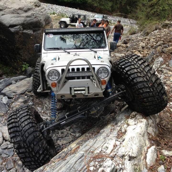 White Jeep with large tires navigating a rocky terrain; people watch in the background.