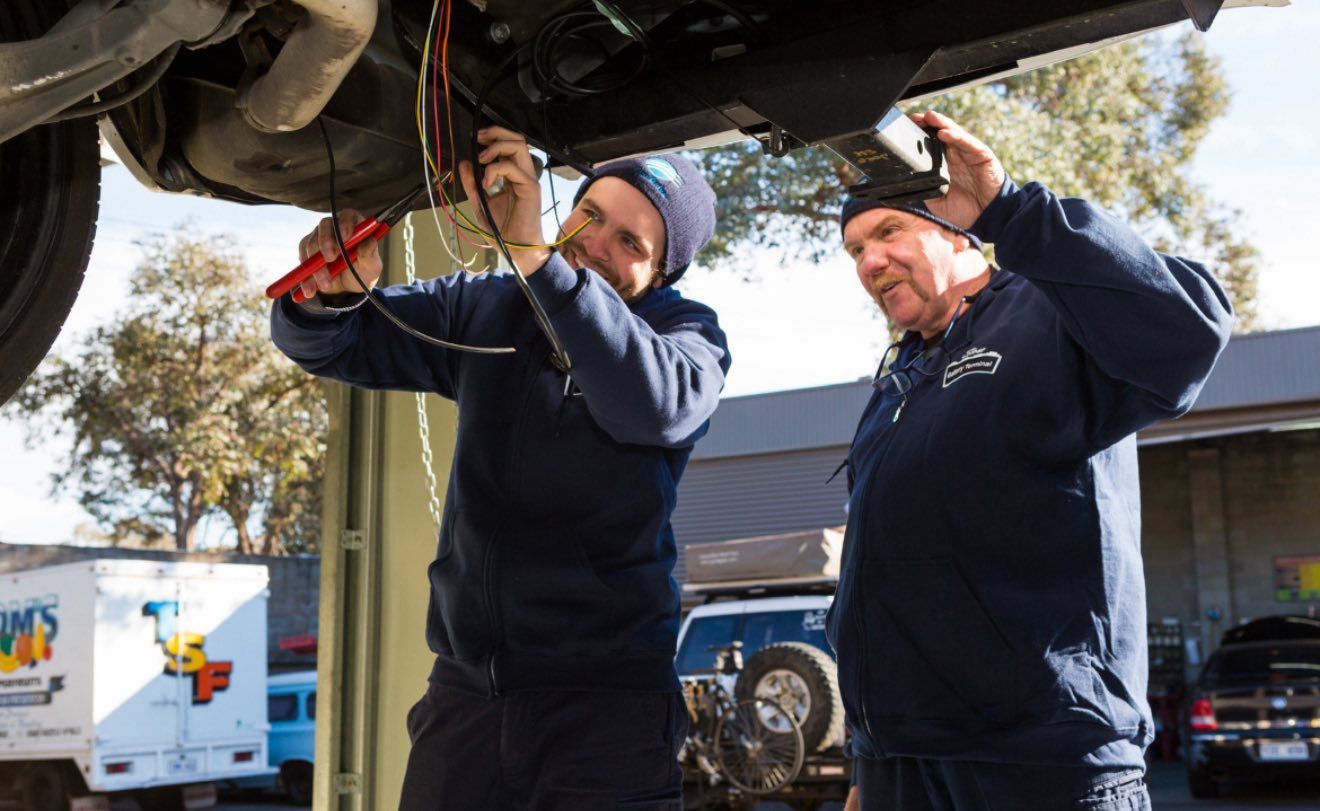Two people working on the underside of a car in a garage, using tools and inspecting wires.
