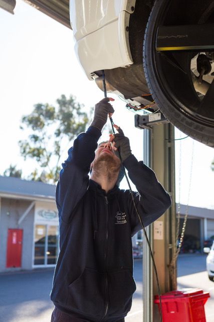 Mechanic works on a car's underside, wearing a blue hoodie and gloves, under a car lift.