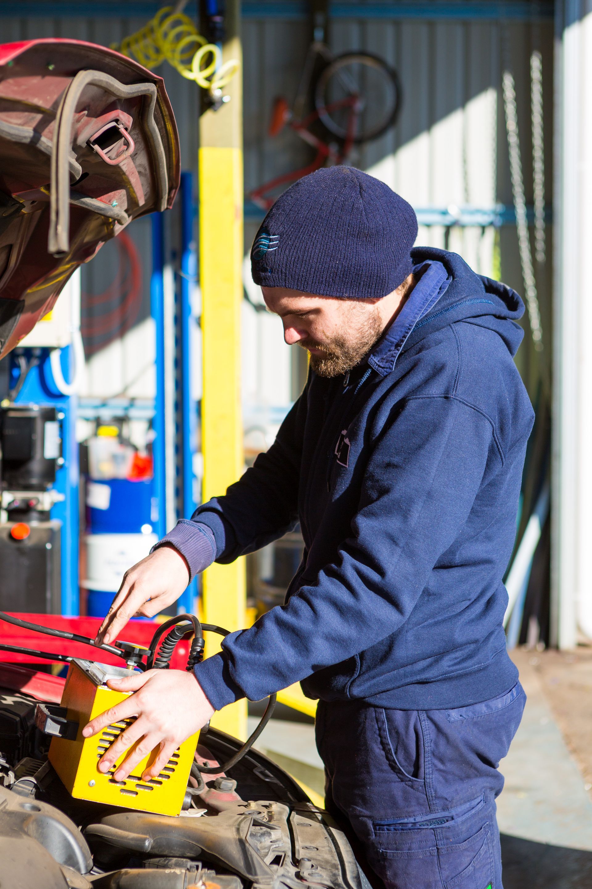 Mechanic in a blue hoodie and beanie charging a car battery in a garage.