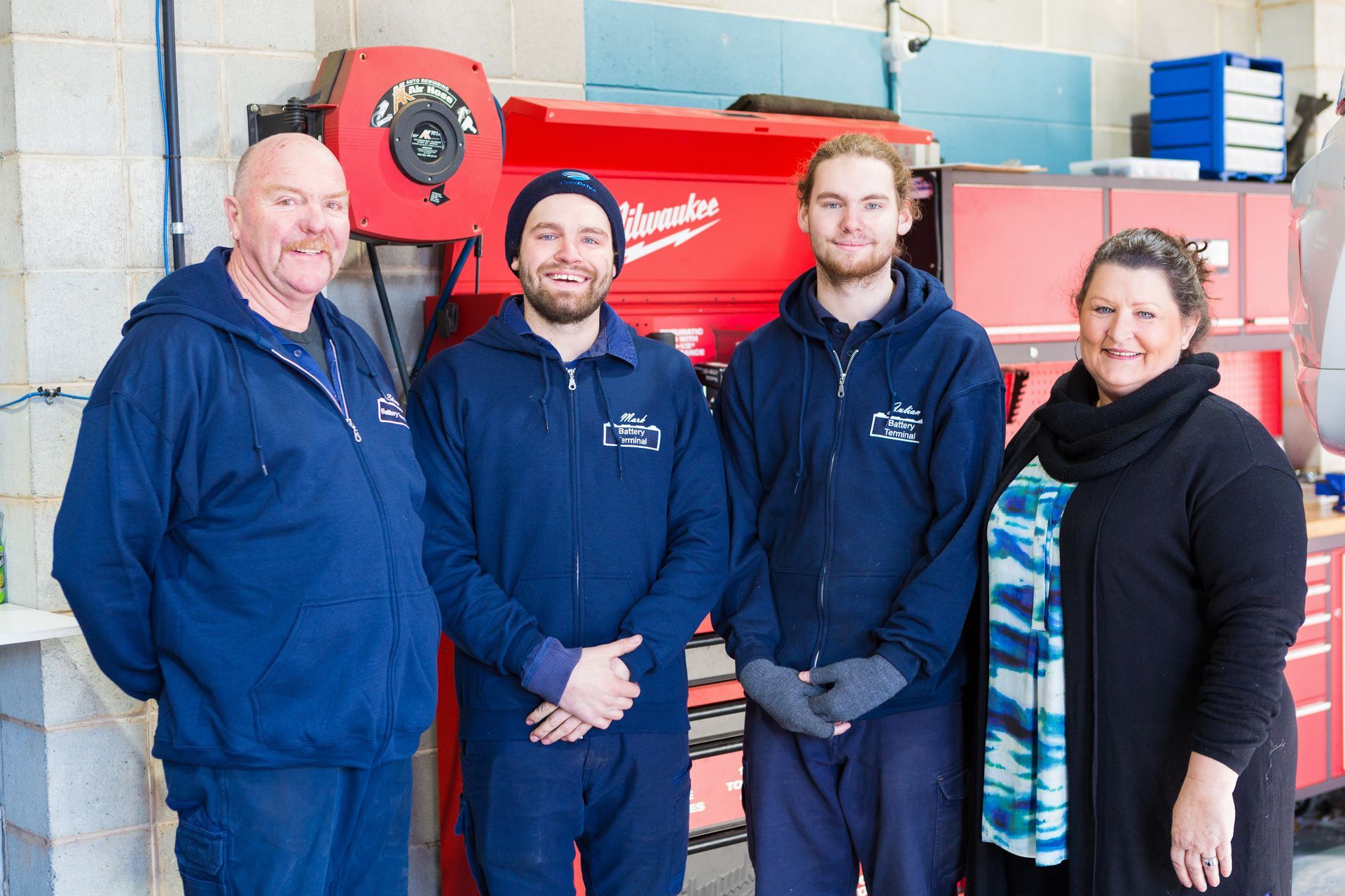Four people in casual attire stand in a workshop with a large red Milwaukee tool chest in the background.