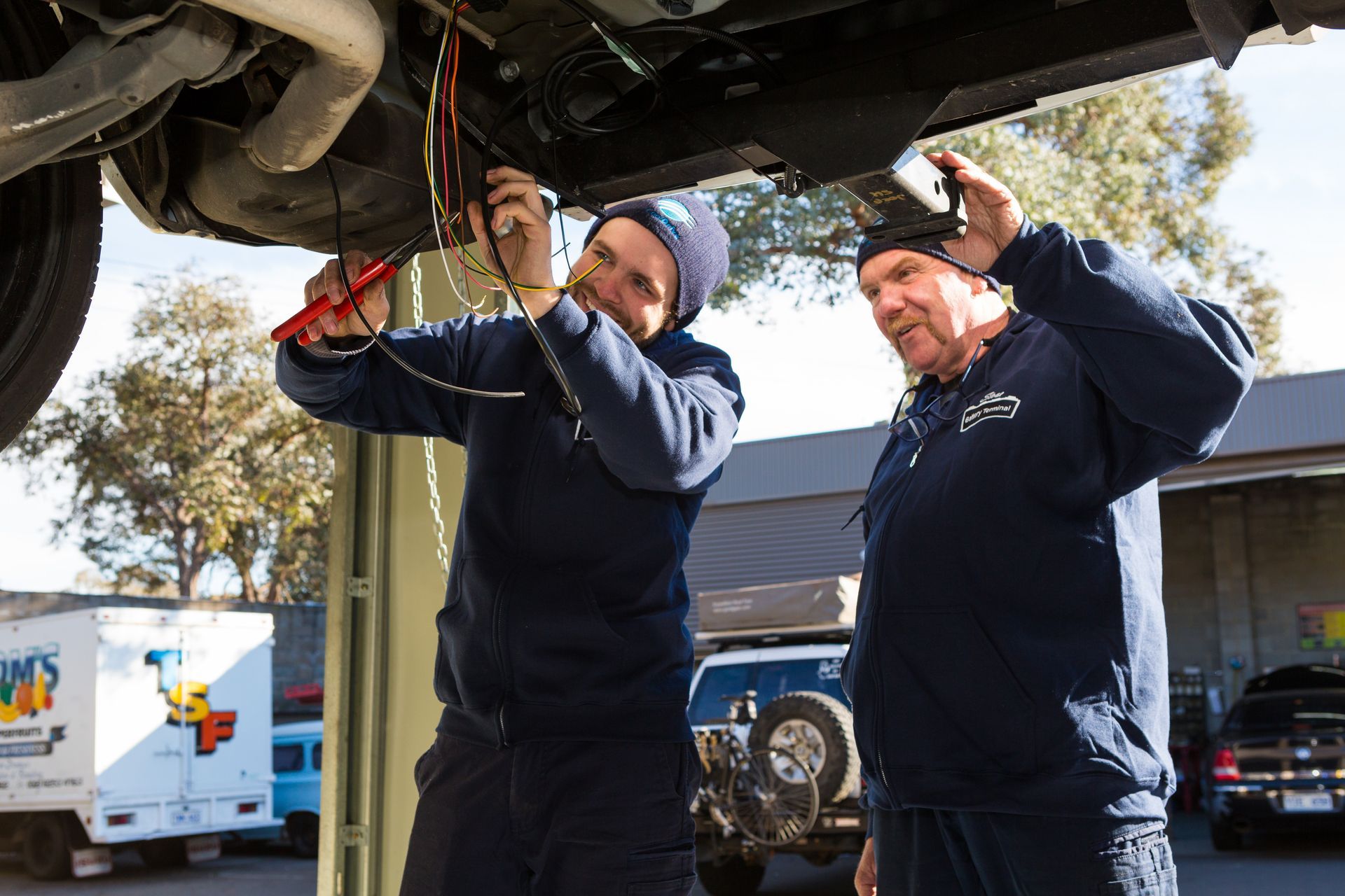 Two mechanics in navy coveralls working under a vehicle in a garage, using tools overhead.