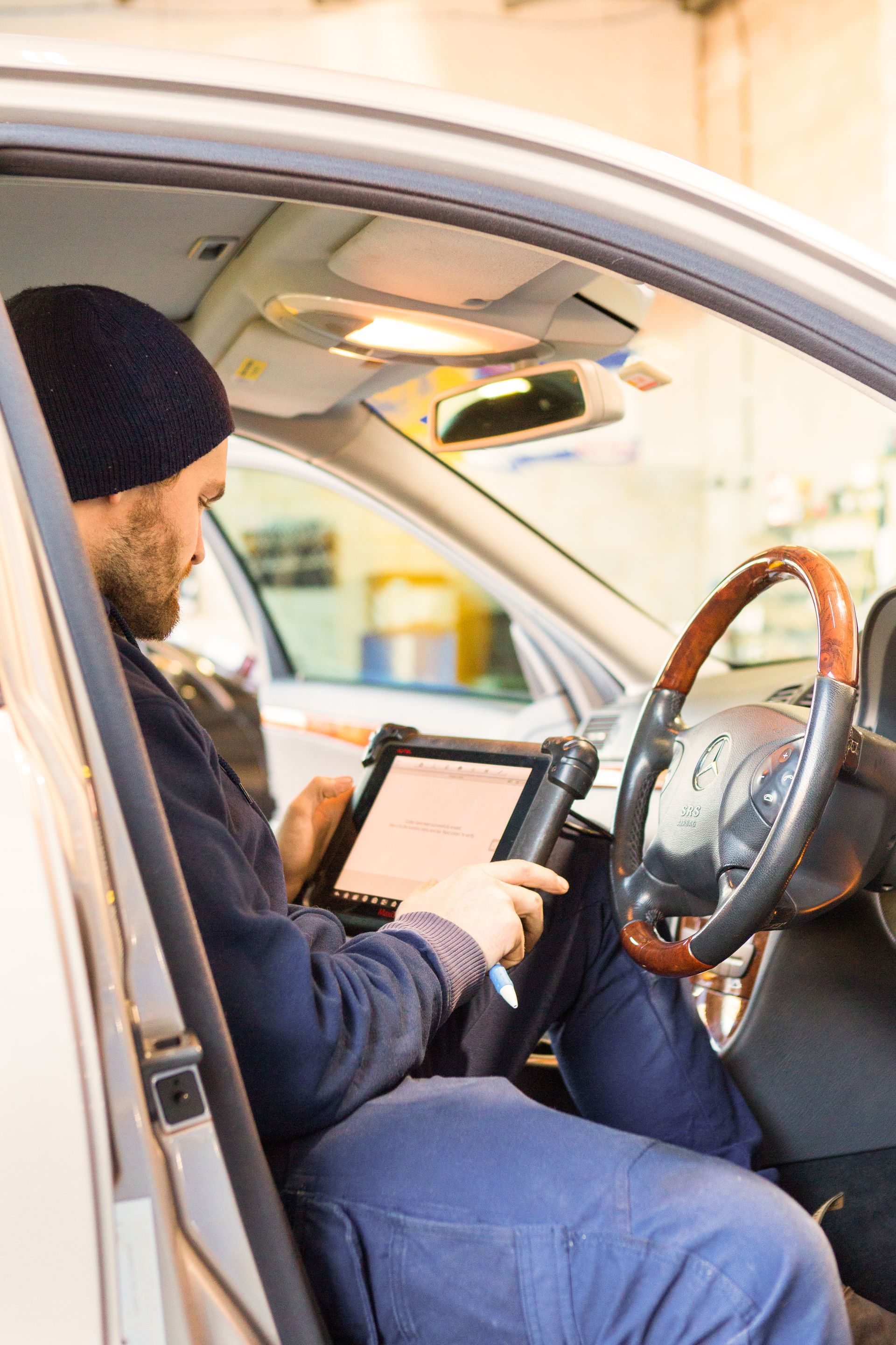 Man in a car using a diagnostic tablet. He’s wearing a beanie and looking at the screen. Sunlight visible.