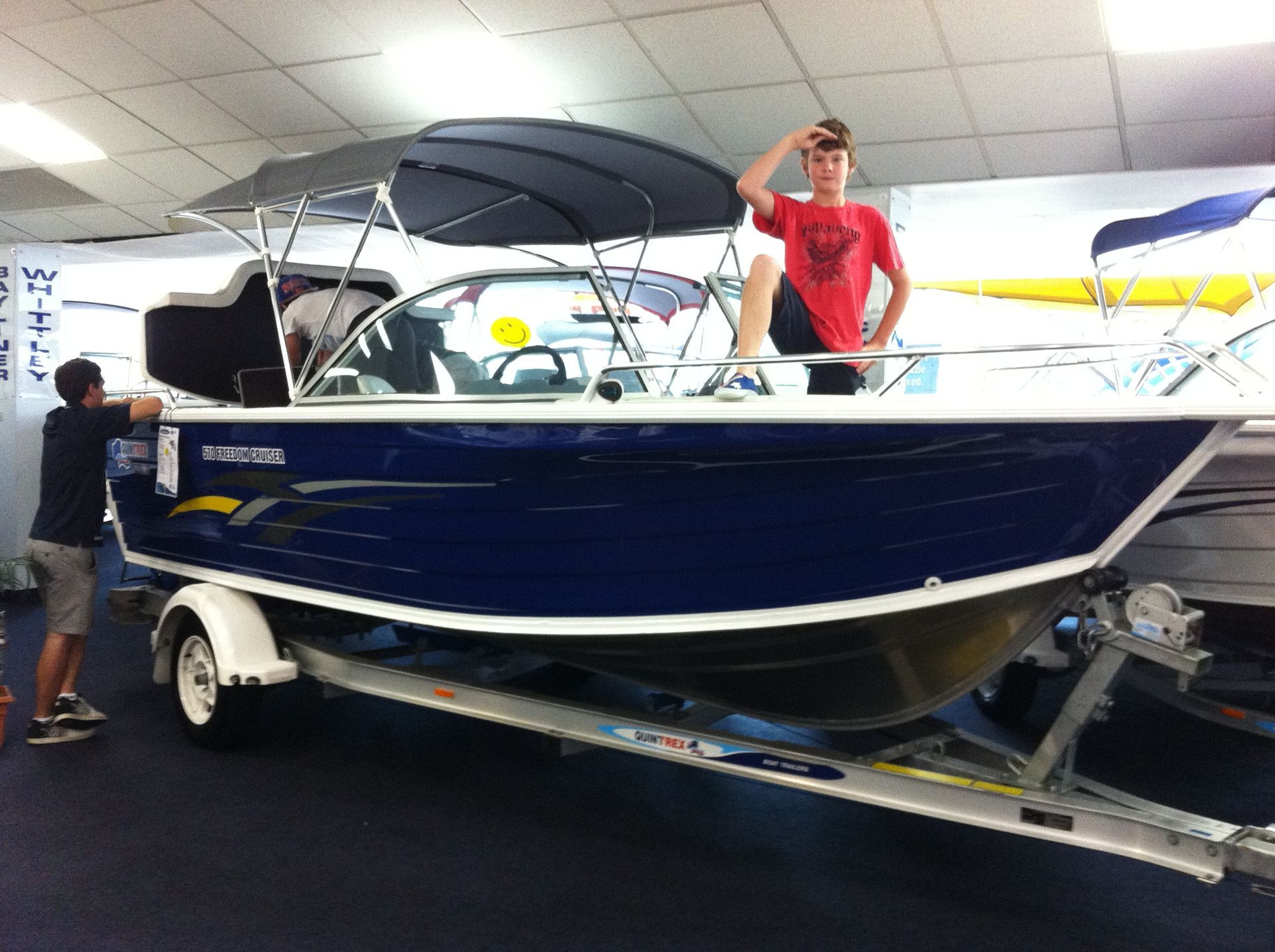 Two people standing near a blue and white boat on a trailer, inside a store. One person salutes while sitting on the boat.