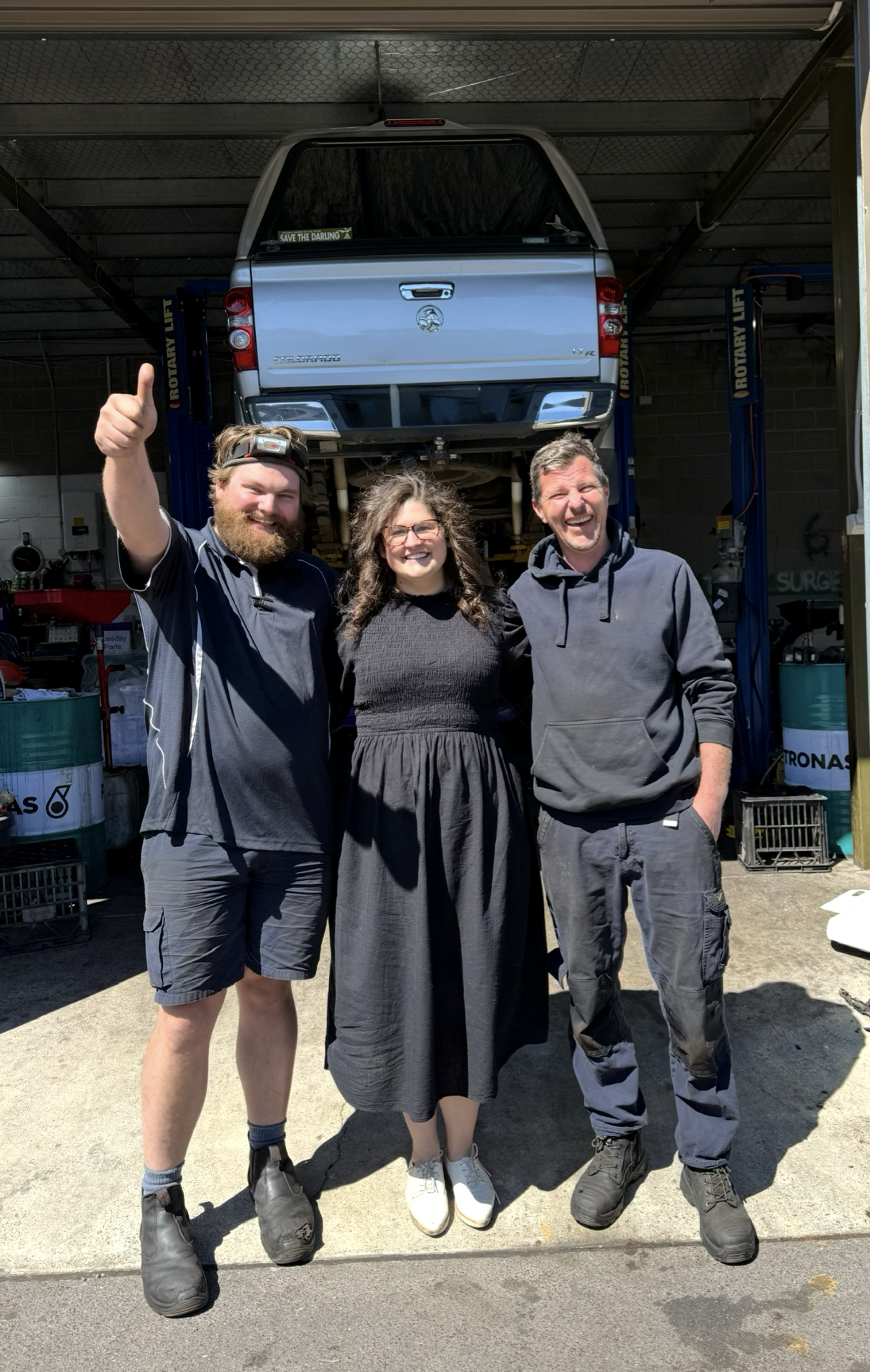 Three people posing in front of a blue car in a garage, one giving a thumbs-up.