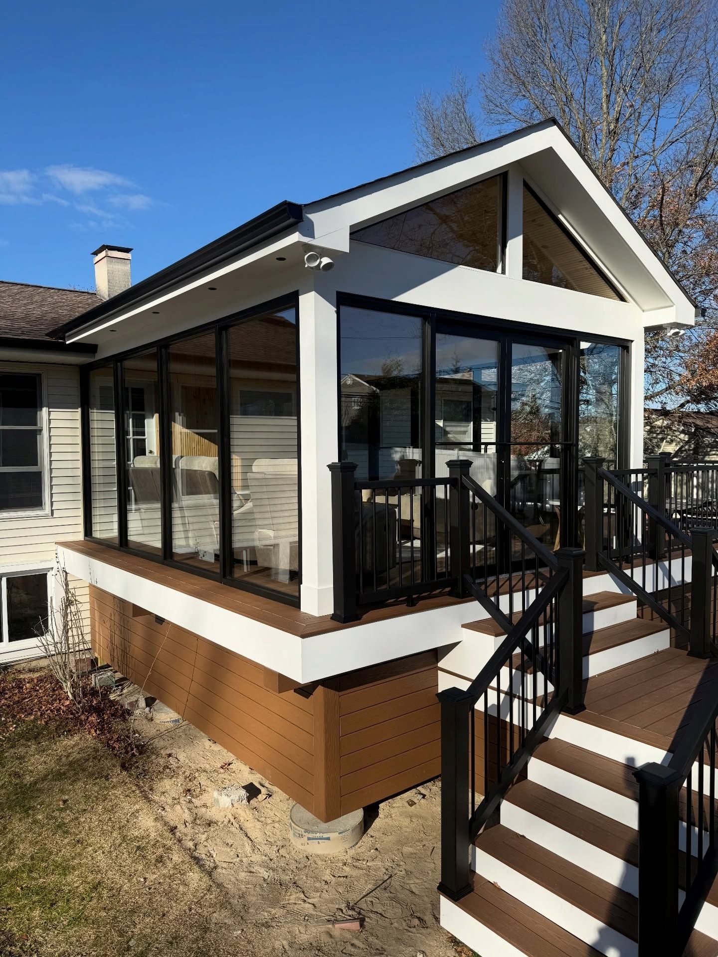 A white-framed sunroom addition with large glass panels, attached to a house with a raised wooden deck and stairs.