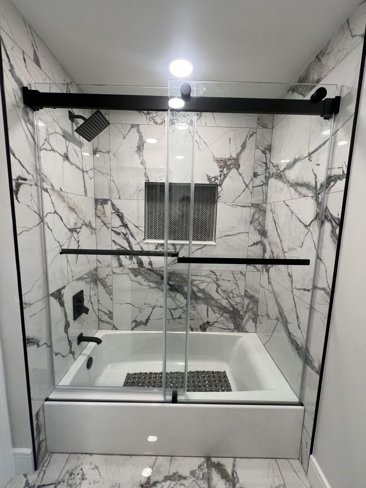 A modern bathtub shower with white marble-patterned tiles, a black sliding glass door, and matte black fixtures.