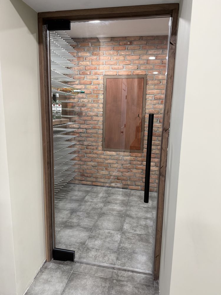 A glass door with a wood frame leads into a wine cellar with brick walls, grey tile flooring, and a small wooden door.