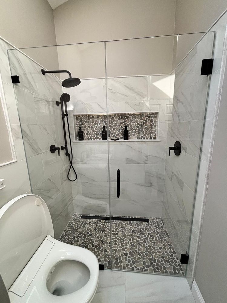 A modern bathroom with a glass-enclosed shower featuring white marble-look tiles, black fixtures, and a pebble-stone floor.