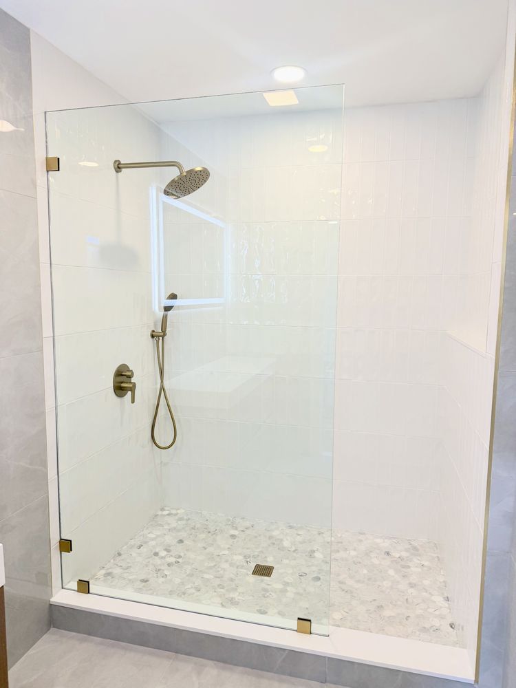 A modern glass-enclosed shower with white tiled walls, pebble flooring, and brushed gold fixtures.