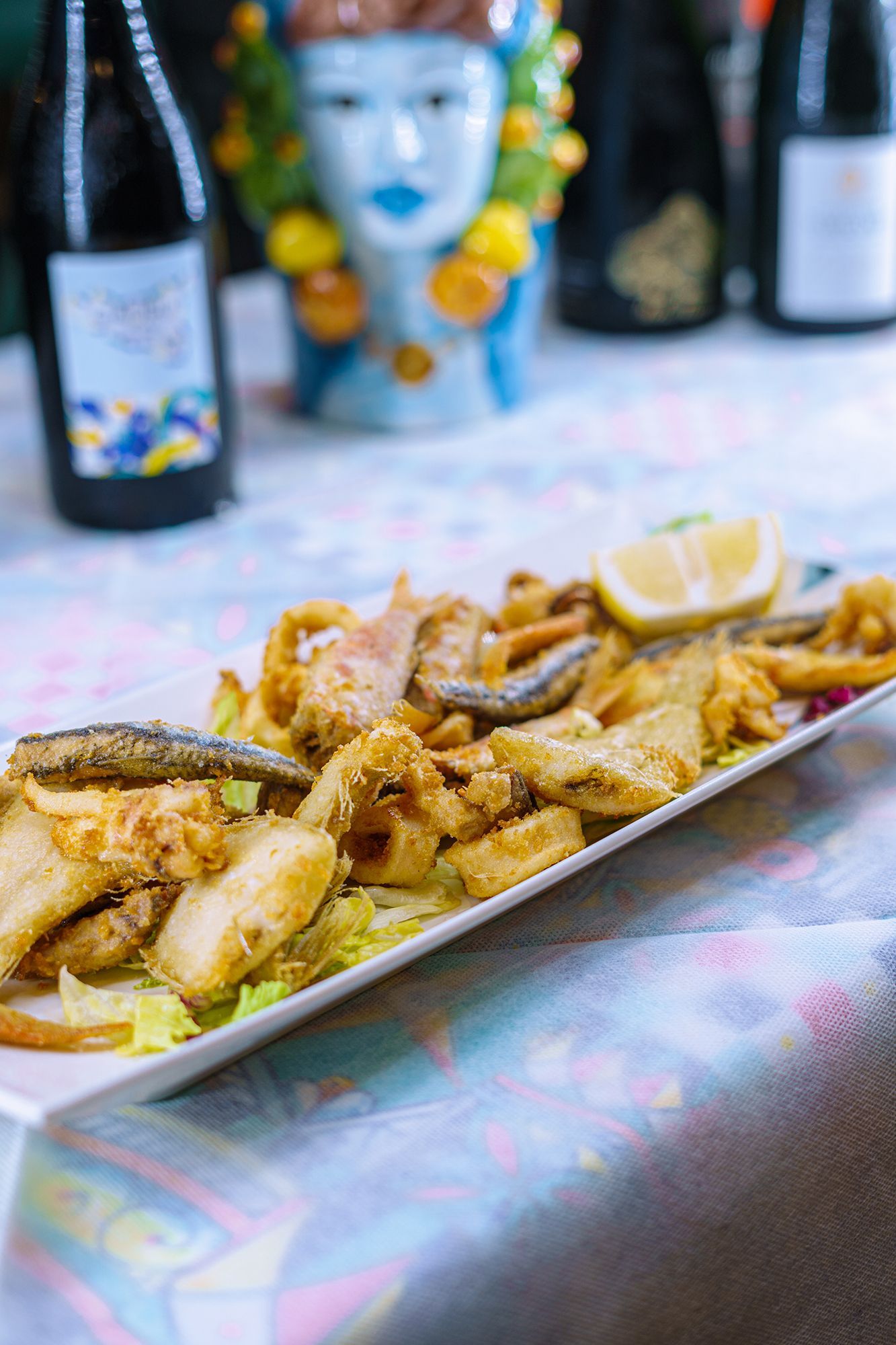 Piatto di pesce fritto con limone su un tavolo con una testa decorativa in ceramica e bottiglie di vino.