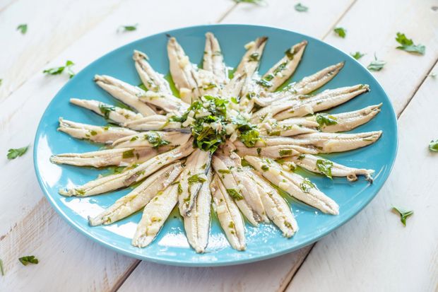 Piatto blu di filetti di pesce bianco ricoperti di erbe aromatiche, disposti a raggiera su un tavolo di legno bianco.