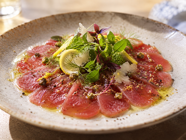 Carpaccio di pesce crudo tagliato a fettine sottili, servito su un piatto con limone, erbe aromatiche e formaggio grattugiato.