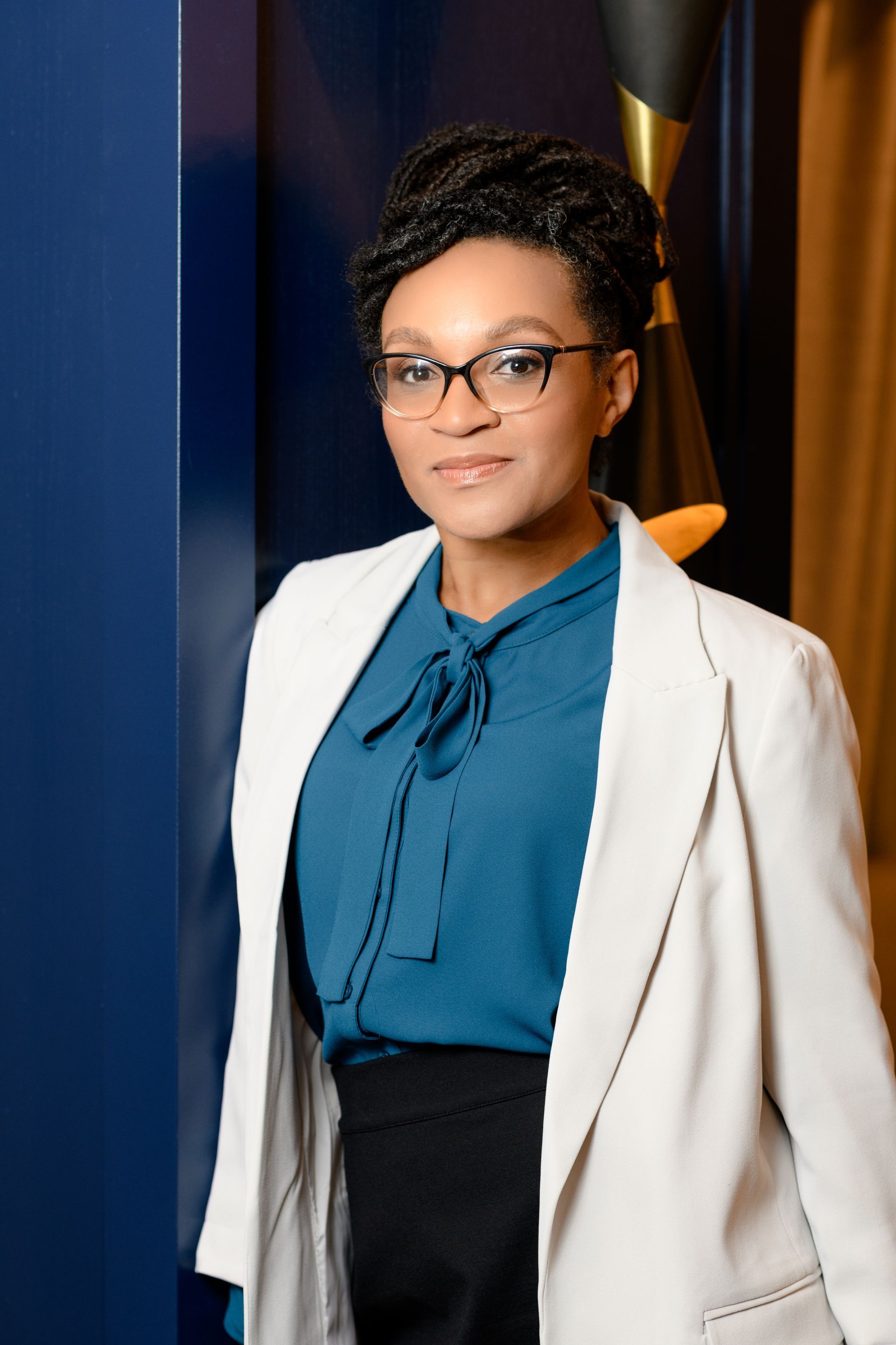 A woman wearing glasses and a white jacket is standing in front of a blue wall.
