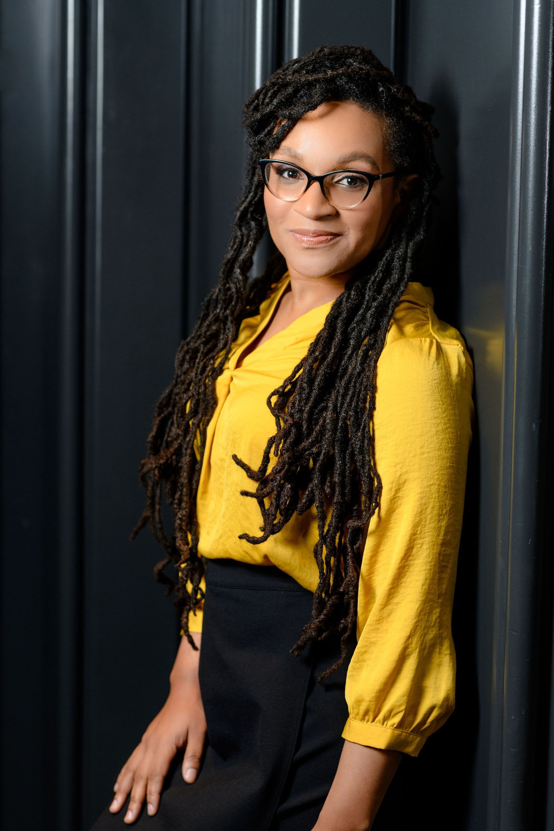 A woman wearing glasses and a yellow shirt is sitting in front of a black wall.