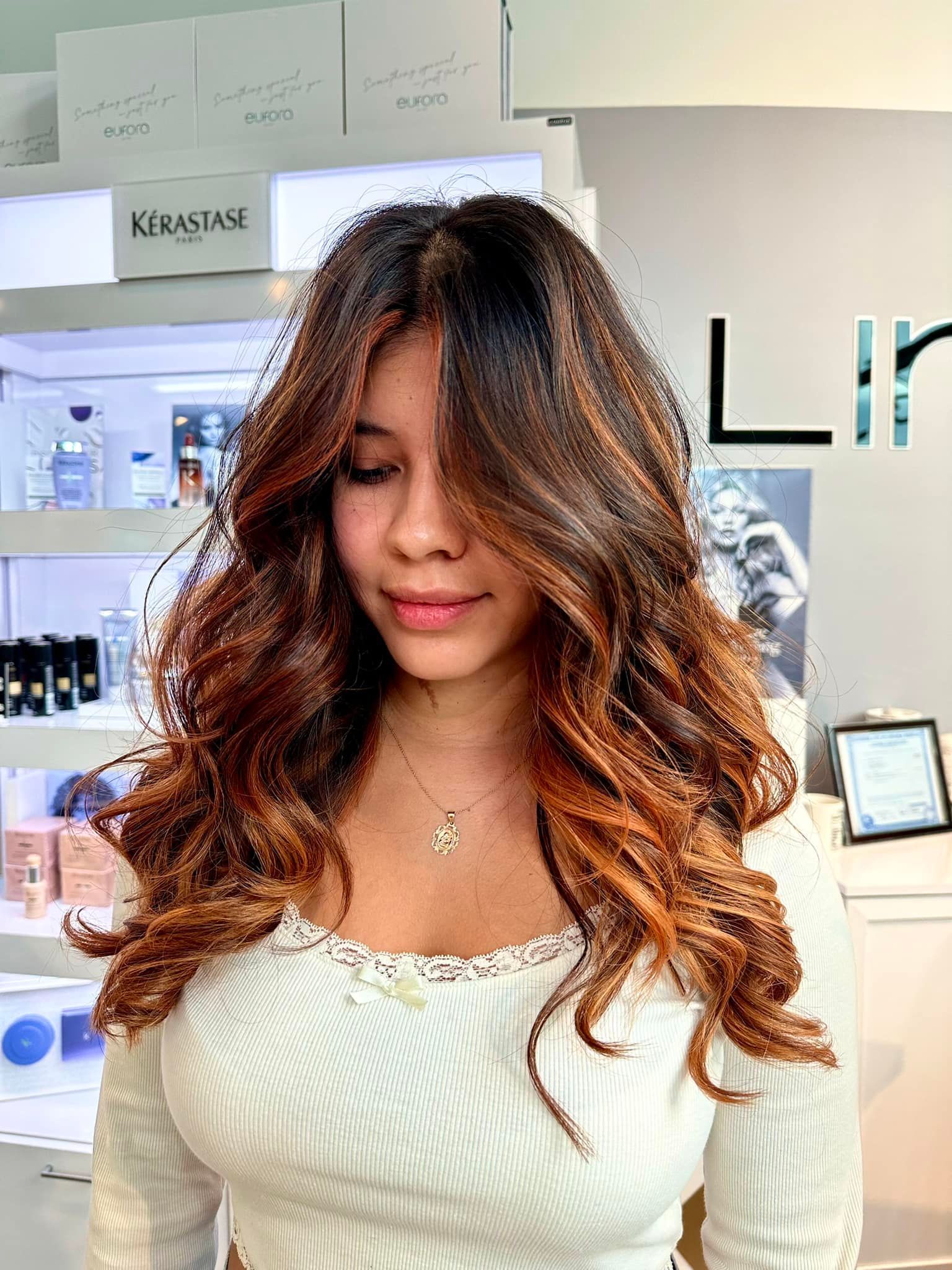 Woman with long, wavy hair, ombre brown to copper. Wearing white top, inside salon with beauty products.