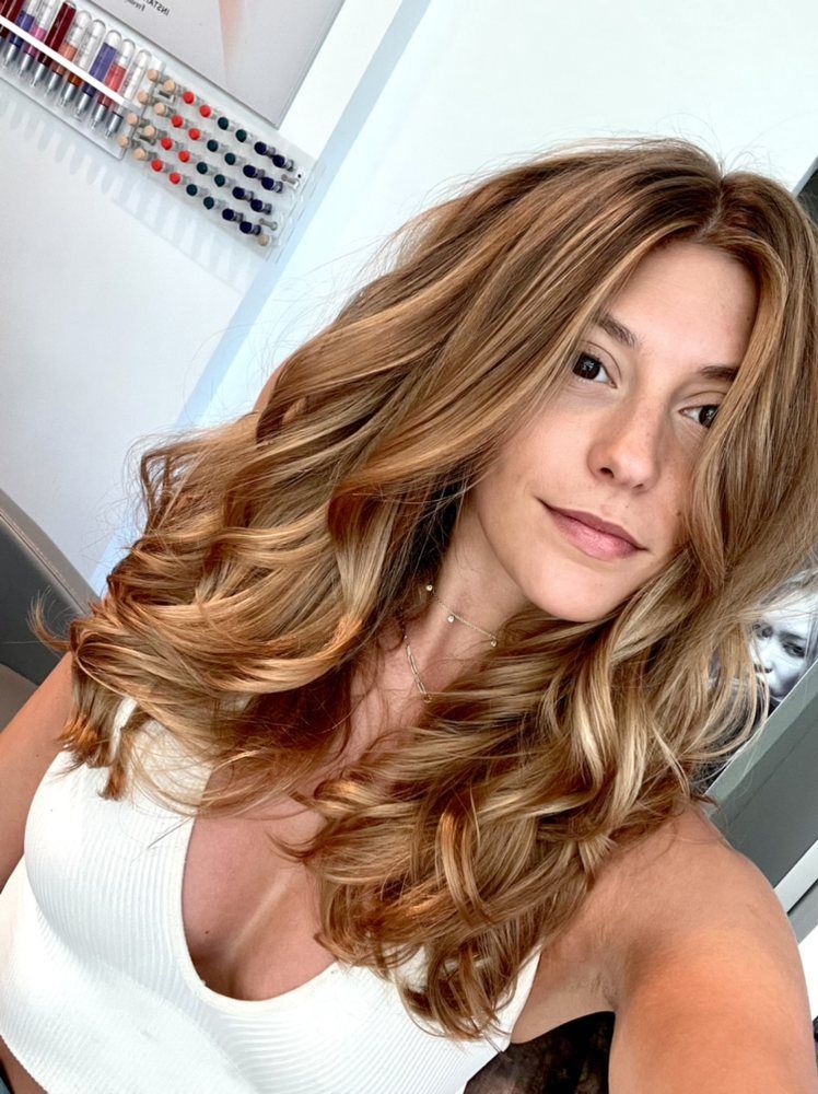 Woman with long, wavy blonde hair after a blow out, wearing a white tank top. Salon setting.